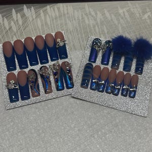 May include: Two sets of artificial nails displayed on silver glittered surfaces. One set features navy blue tips with clear rhinestones, while the other showcases a variety of designs, including blue and silver accents, and fluffy blue pom-poms.