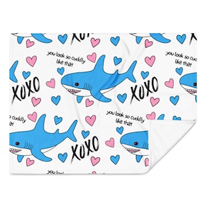 May include: White blanket with a repeating pattern of blue sharks, pink and blue hearts, and the text "XOXO" and "you look so cuddly like that". The blanket is folded over, showing a white underside.