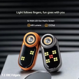 May include: Two small, handheld devices with a metallic finish. One is orange, the other silver. Each has a central light and a screen displaying a grid of illuminated squares. Text on the image reads: "Light follows fingers, fun goes with you."