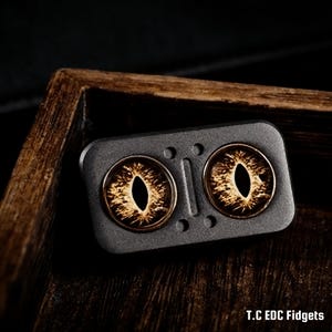 Pode incluir: Um fidget spinner cinza escuro com dois desenhos em forma de olho. Os olhos são castanhos e dourados com uma pupila oval preta. O spinner está sobre uma superfície de madeira. O texto no canto inferior direito diz "T.C EDC Fidgets."