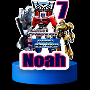 Pode incluir: Um topo de bolo azul e branco com os personagens Transformers Optimus Prime, Bumblebee e Megatron. O topo também inclui o texto "Transformers", "Feliz aniversário" e "Noah" em uma fonte rosa e azul.
