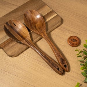 May include: Two wooden salad spoons and a wooden coaster are displayed on a wooden cutting board. The spoons are engraved with the names "Michael" and "Amelia". The coaster has the text "Est. 2023" and a design.