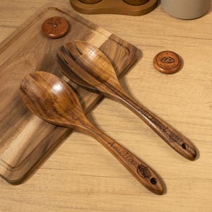 May include: Wooden salad spoon and fork set with engraved names and decorative details. The set includes a solid spoon and a slotted fork, both crafted from a rich, dark wood. The set rests on a wooden cutting board.