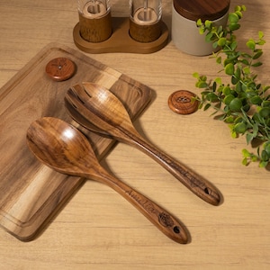 May include: Two wooden salad spoons with the name "Amelia" engraved on one and "Wild Sweet" on the other. The spoons are on a wooden cutting board. Other wooden kitchen accessories are in the background.
