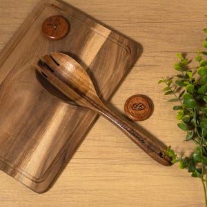 May include: Wooden salad spoon with a slotted head and a cutting board. The spoon has the name "Michael" engraved on the handle. Two small wooden coasters are also present, one with a floral design and the other with a bread design.