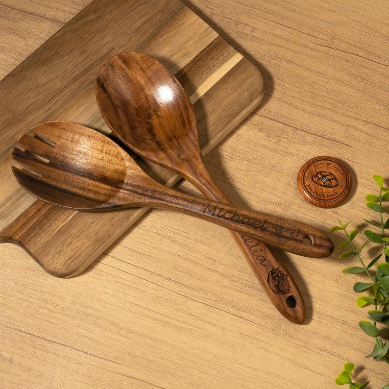 May include: Wooden salad serving spoons with engraved names and a small wooden coaster. The spoons are brown with a smooth finish, and one has slots for draining. The coaster has a bread design.
