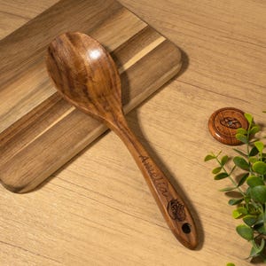 May include: A wooden spoon with the name "Amelia" engraved on the handle. The spoon is made of a light brown wood and has a round bowl. A small wooden coaster and greenery are also in the image.