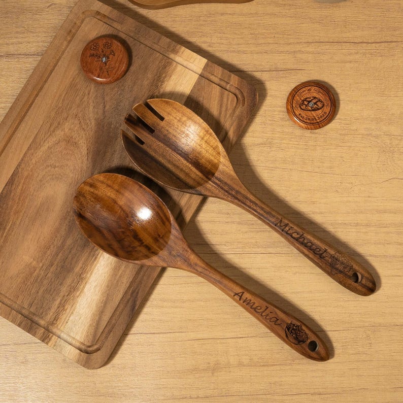 May include: Two wooden salad spoons and two wooden coasters rest on a wooden cutting board. One spoon is solid, the other has slots. The spoons are engraved with the names "Amelia" and "Michael". The coasters have engraved designs.