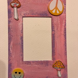 May include: A rectangular picture frame painted in pink and purple with a white center. The frame is decorated with a peace sign, a smiley face, and two mushroom designs. The frame has a textured, painted appearance.