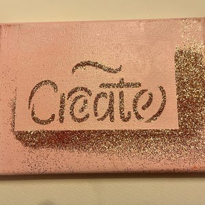 May include: A pink canvas with the word "Creative" written in gold glitter. The word is surrounded by a gold glitter border.