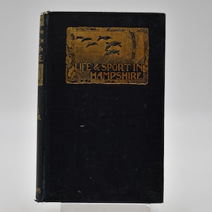 Pode incluir: Um livro azul escuro com um título dourado que diz "Life & Sport in Hampshire". O título é rodeado por uma borda decorativa com um desenho de pássaros em voo.