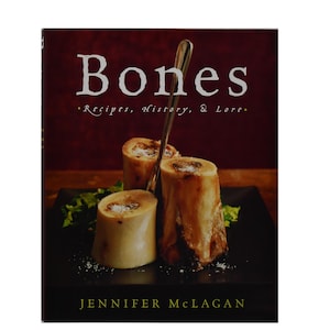 May include: A cookbook titled "Bones" with the subtitle "Recipes, History, & Lore." The cover features three roasted bone marrow pieces on a black plate, garnished with greens. A silver serving utensil is inserted into one bone. The author's name, Jennifer McLagan, is at the bottom.