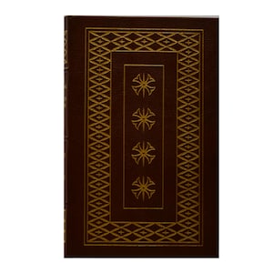 Könnte beinhalten: Ein Hardcover-Buch mit dunkelbraunem Einband und goldenen Zierakzenten. Der Einband zeigt ein rechteckiges Design mit geometrischen Mustern und vier stilisierten floralen Elementen. Der Buchrücken ist ebenfalls dunkelbraun.