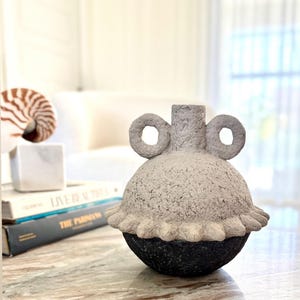 Pode incluir: Um vaso decorativo com um corpo texturizado cinza claro e uma base preta. O vaso tem duas alças circulares e uma borda com babados. O vaso está sobre uma superfície com padrão de mármore.