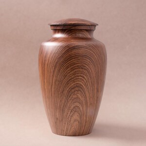 May include: A wooden urn with a dark brown wood grain pattern and a lid. The urn is a traditional shape with a rounded body and a narrow neck.