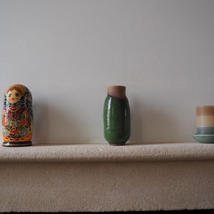 Peut inclure: Une poupée russe colorée, un vase vert avec un dessus marron et une bougie superposée sur une petite soucoupe sont exposés sur une étagère en pierre. La poupée a un visage peint et des motifs floraux. La bougie a des couches beiges, pêche et bleues.