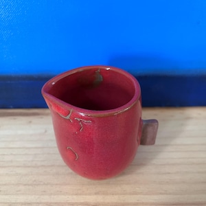 Peut inclure: Un petit pot à crème en céramique rouge avec une poignée. Le pot à crème a une forme arrondie et un bord légèrement irrégulier. La poignée est de forme rectangulaire. Le pot à crème est sur une surface en bois avec un fond bleu.