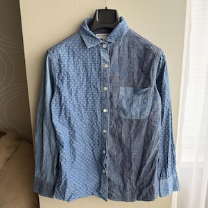 May include: A blue denim shirt with long sleeves and a button-down front. The shirt features a unique design with a textured pattern on the left side and a solid colour on the right side, along with a chest pocket.