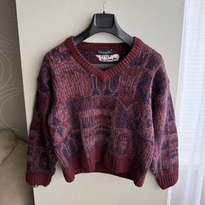 Può includere: Un maglione bordeaux con scollo a V con un motivo geometrico blu navy e bordeaux. Il maglione è appeso a una gruccia nera. L'etichetta recita "Christian Dior".