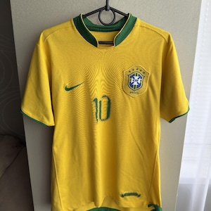 Puede incluir: Una camiseta de fútbol amarilla con ribetes verdes y el número 10 en verde. La camiseta presenta el emblema de la bandera brasileña y un logotipo de Nike. La camiseta está colgada de una percha negra.