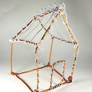 May include: A wire house sculpture made of copper and silver wire. The house has a simple rectangular shape with a roof and a door. The wire is twisted and curled to create a decorative effect.