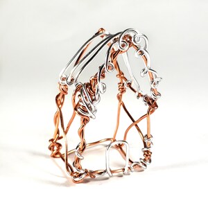 May include: A small, wire-framed house sculpture made of copper and silver wire. The house has a simple, rectangular shape with a pointed roof and a small door.
