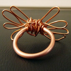 May include: A copper wire ring with a decorative design of multiple loops and swirls. The ring is made of a single piece of wire that is twisted and shaped to create the design.