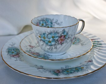 Könnte beinhalten: Ein weißes Tee-Set mit Untertasse, das mit einem Blumenmuster mit blauen und rosa Blüten verziert ist. Das Set hat einen goldenen Rand.