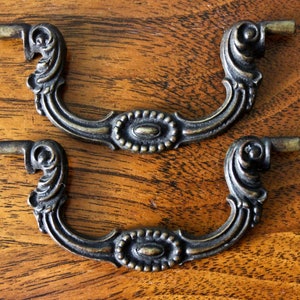 Può includere: Due tiretti per cassetti in metallo decorati con un design ornamentale. I tiretti sono di colore ottone scuro e hanno una forma curva con un ovale centrale.