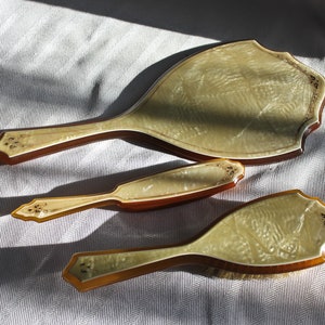 Può includere: Un set da toeletta vintage con uno specchio da mano, un pettine e una spazzola. Il set è realizzato in un materiale marrone chiaro con una finitura bianca perlata. I manici sono decorati con motivi floreali.