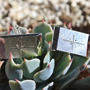 May include: A pair of silver cufflinks with a starburst design. The cufflinks are sitting on a green succulent plant.