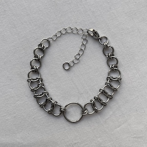 Puede incluir: Una pulsera de cadena en tono plateado construida con anillos metálicos circulares entrelazados. El diseño presenta un anillo central más grande conectado a eslabones repetitivos más pequeños a ambos lados, con un cierre de mosquetón y una cadena extensora ajustable.