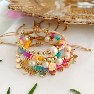 May include: A stack of colorful beaded bracelets with gold accents and pearl embellishments. The bracelets feature a variety of colors, including pink, yellow, turquoise, and white. One bracelet has a gold charm with a smiling face.