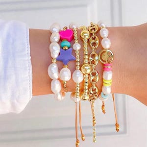 May include: A collection of pearl and gold bracelets. One bracelet features a pink heart charm, a turquoise bead, and a purple star charm. Other bracelets have gold chain links and colorful beads. The bracelets are on a person's wrist.