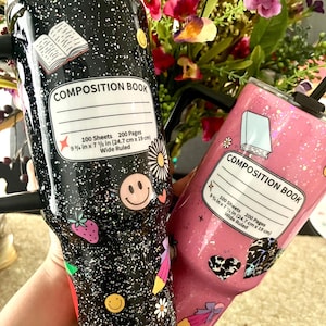 May include: Two stainless steel tumblers with glitter and colorful designs. The black tumbler has a composition book design with the text "COMPOSITION BOOK 100 Sheets 200 Pages 9 1/4 in x 7 1/2 in (24.7 cm x 19 cm) Wide Ruled". The pink tumbler has a similar design with the text "COMPOSITION BOOK 100 Sheets 200 Pages 9 1/4 in x 7 1/2 in (24.7 cm x 19 cm) Wide Ruled".