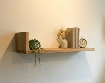 Può includere: Una mensola galleggiante in legno con una piccola pianta in vaso, un vaso con fiori secchi, libri e un orologio decorativo. La mensola è in legno chiaro ed è montata su una parete di colore neutro. La mensola è una soluzione di archiviazione decorativa.