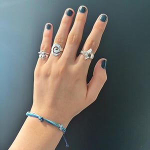 Può includere: Una mano con quattro anelli d'argento, uno con un design a spirale, uno con una forma geometrica e due bande semplici. La mano indossa anche un braccialetto dell'amicizia blu e bianco.