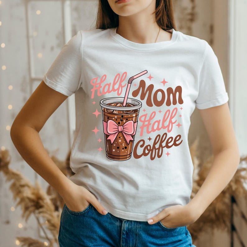 Pode incluir: T-shirt branco com a frase "Half Mom Half Coffee" em rosa e castanho. O design inclui uma ch&aacute;vena de caf&eacute; com um la&ccedil;o rosa e um canudo. Uma pe&ccedil;a de roupa casual para o dia a dia.