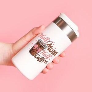 Pode incluir: Caneca de viagem branca com aro de a&ccedil;o inoxid&aacute;vel e tampa branca. A caneca apresenta um gr&aacute;fico de uma bebida de caf&eacute; com um la&ccedil;o rosa e as palavras "Half Mom Half Coffee". O fundo &eacute; rosa.