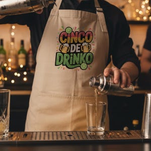 May include: A beige apron with the text "Cinco de Drinko" in colorful lettering. The apron has a pocket and is being worn by a person pouring a drink from a cocktail shaker into a glass. The background includes bar equipment.