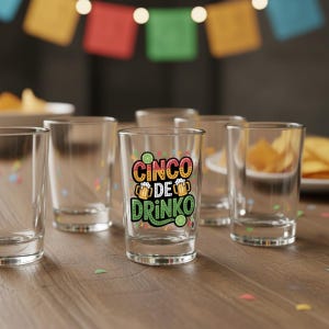 May include: Clear shot glasses with the text "Cinco de Drinko" in colorful lettering. The shot glasses are arranged on a wooden table, with colorful confetti and a festive banner in the background. A plate of chips is visible.