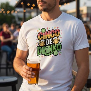 May include: White t-shirt with the text "CINCO DE DRINKO" in colorful lettering, featuring beer mugs and lime slices. The person in the image is holding a pint glass of beer. The shirt is a casual style.