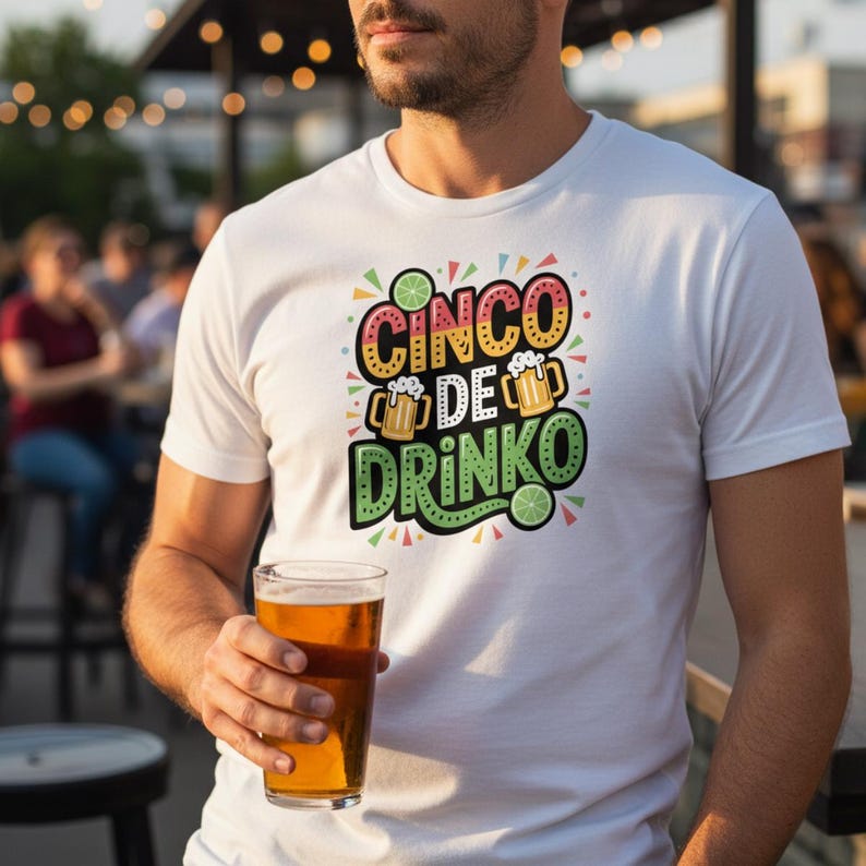 May include: White t-shirt with the text "CINCO DE DRINKO" in colorful lettering, featuring beer mugs and lime slices. The person in the image is holding a pint glass of beer. The shirt is a casual style.