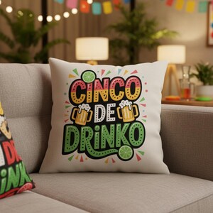 May include: A square, off-white throw pillow with the words "CINCO DE DRINKO" in colorful, block letters. The design includes images of beer mugs and lime slices, perfect for a festive celebration. The pillow is resting on a gray sofa.