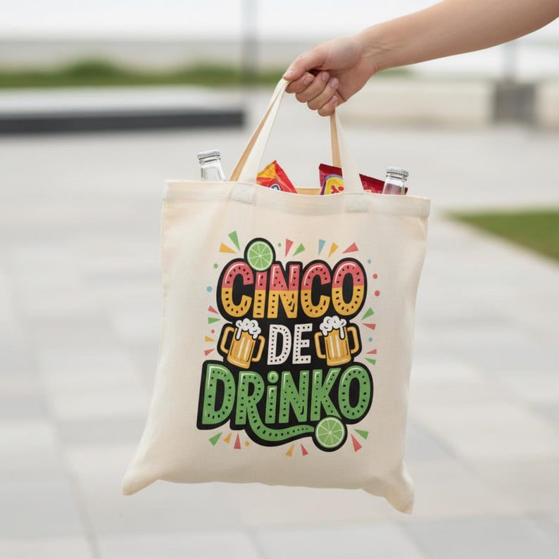 May include: A beige canvas tote bag with the words "Cinco de Drinko" in colorful lettering. The design includes beer mugs and lime slices. The bag is filled with snacks and drinks and is held by a person.