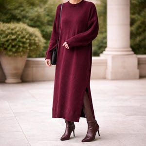 May include: A long burgundy sweater dress with a crew neck and side slits. The dress is worn with matching burgundy heeled boots and a black shoulder bag. The outfit is suitable for cooler weather.