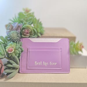May include: A lavender cardholder with the text "Best life Ever" in silver lettering. The cardholder has a pocket on the front and is surrounded by green and purple succulents.