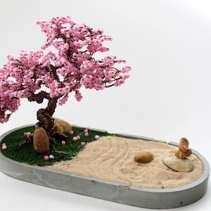 Op de afbeelding: Een roze kralenbonsaiboom met een grijze betonnen basis en een met zand gevulde lade. De boom staat in een kleine, ovale lade met een groene mosbasis en kleine stenen.