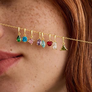 May include: A gold chain necklace featuring a series of small, dangling charms with multi-colored gemstones in various shapes, including pear, round, square, marquise, heart, rectangular, and triangular cuts, with the text NikkiMinimalCo.