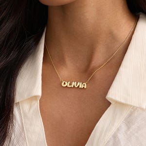 May include: A gold-toned metal necklace featuring a nameplate pendant with the name OLIVIA in a rounded, bubbly font. The pendant is attached to a delicate, thin gold-toned chain that rests against the collarbone area of the neck.
